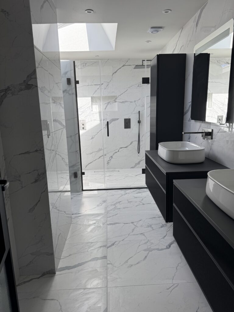 Modern bathroom with white marble walls and floors, a large glass-enclosed shower with black fixtures, black vanities with vessel sinks, and a skylight letting in natural light.