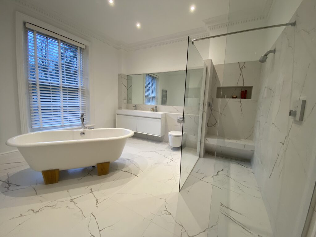 Spacious modern bathroom with white marble floors and walls, a freestanding bathtub, large mirror, floating vanity, glass-enclosed shower, and a large window with white blinds.