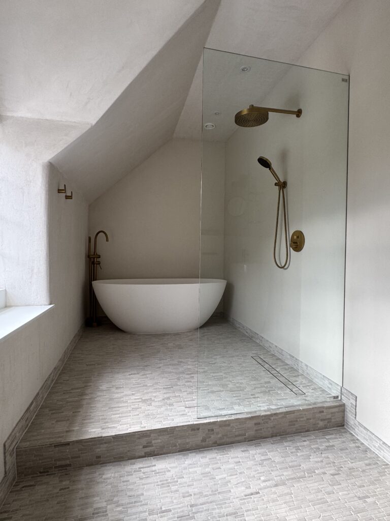 Modern bathroom with a freestanding white bathtub, gold fixtures, a walk-in shower with a glass partition, sloped ceiling, and light gray tiled floor. The space is minimalist and bright.