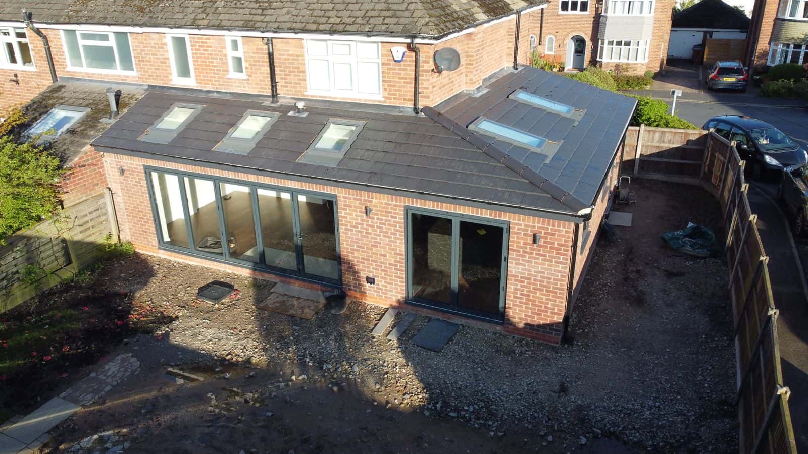 Aerial view of a brick house extension with large glass doors and several skylights on the roof, surrounded by a bare, unfinished yard. The extension is attached to a two-story brick home in a suburban neighborhood.
