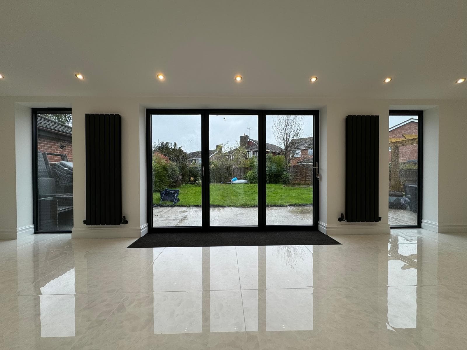 A bright, modern room with glossy tiled floors and large black-framed glass doors overlooking a green garden. Two tall black radiators are mounted on either side of the doors.