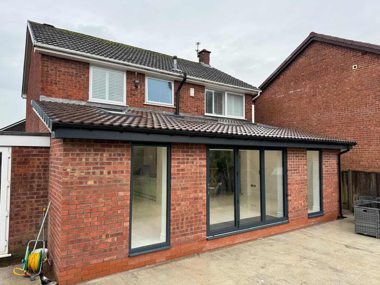 A modern red-brick house with a new extension featuring large floor-to-ceiling windows and a tiled roof. A garden hose is coiled on the ground outside on a concrete patio. The sky is overcast.