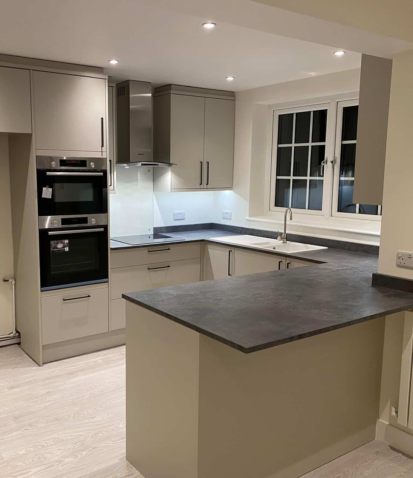 Modern kitchen with beige cabinets, black countertops, built-in oven and microwave, an induction cooktop with hood, a sink under a window, and light wood flooring. The space is well-lit with recessed ceiling lights.