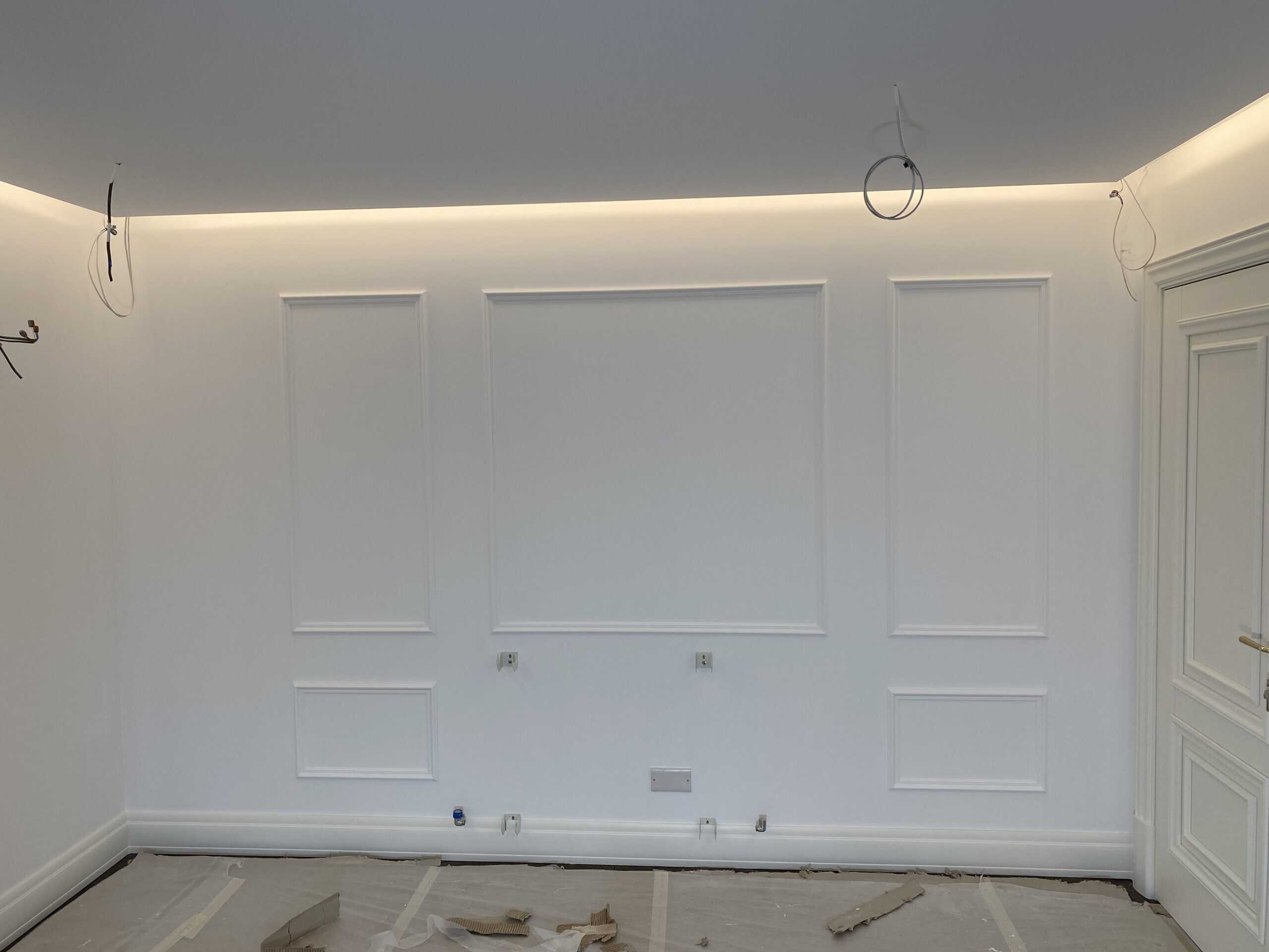 A room with white walls featuring decorative moulding. The ceiling has recessed lighting, and electrical wires are protruding. The floor is covered with protective sheets and some scattered debris. A white door is on the right.