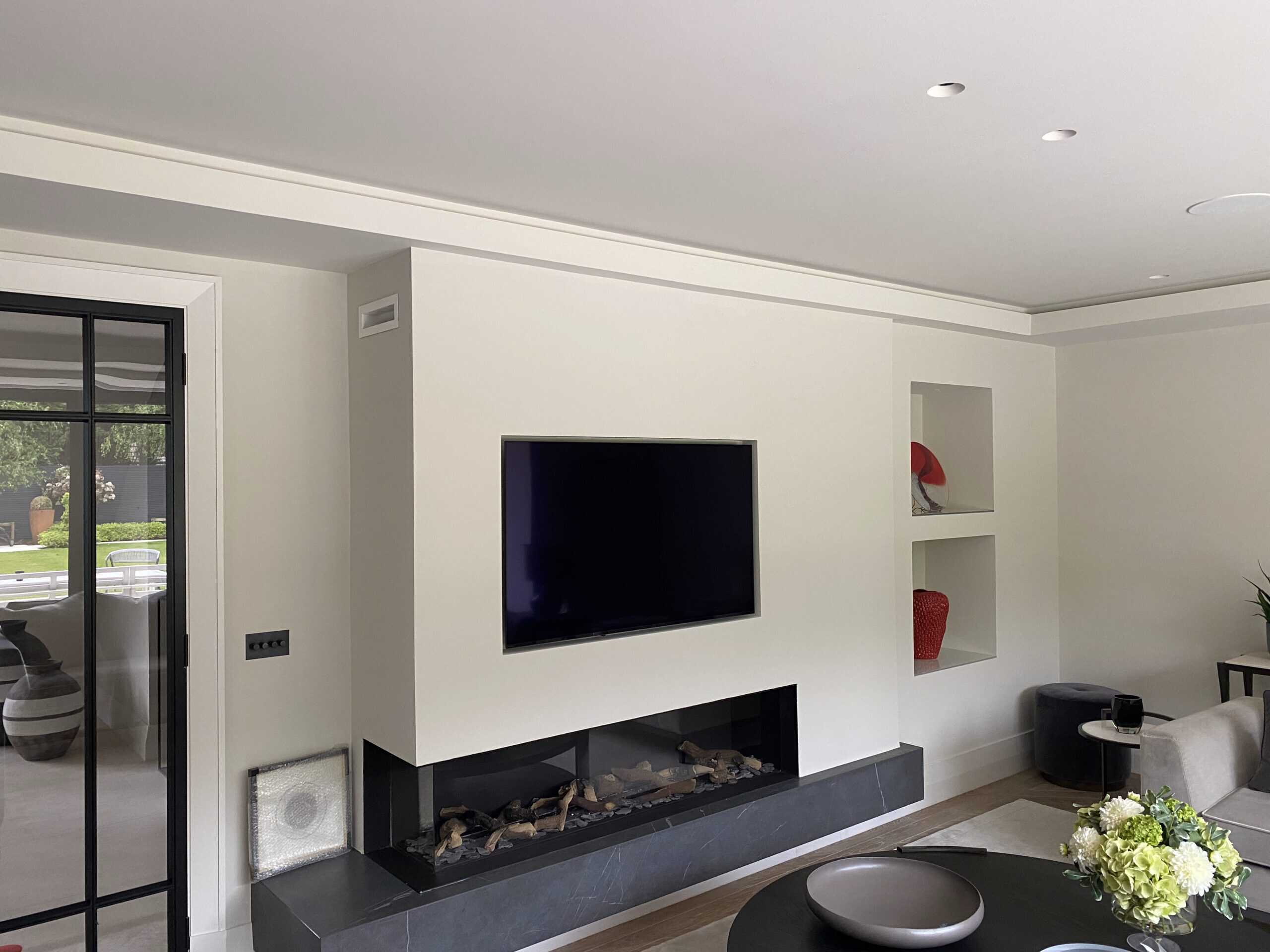 A modern living room with a wall-mounted TV above a sleek fireplace. The room features minimalist decor, including built-in shelves with red vases and a decorative bowl on a table. A large window offers a view of greenery outside.