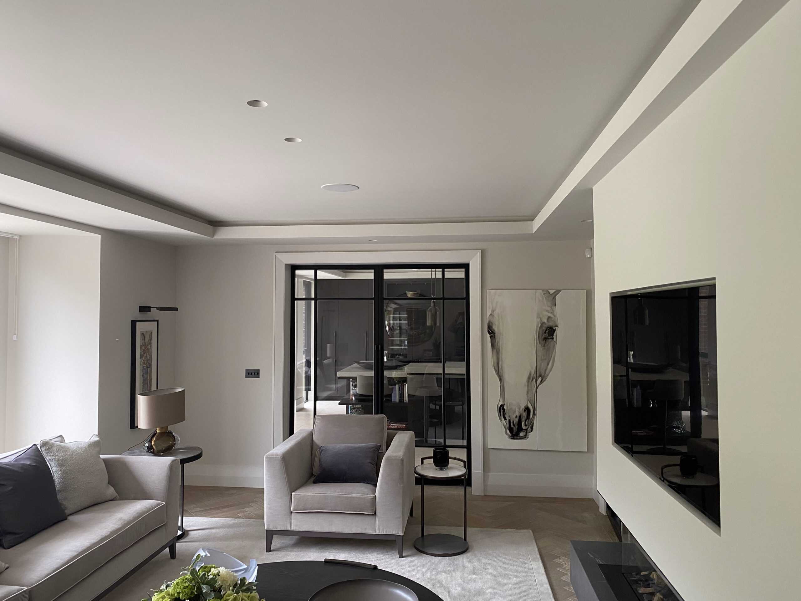 Modern living room with a large painting of a horse's face on the wall. The space features a beige armchair, sofa, side table with a lamp, and a wall-mounted flat-screen TV. Neutral tones and a glass door create a calm, elegant ambiance.