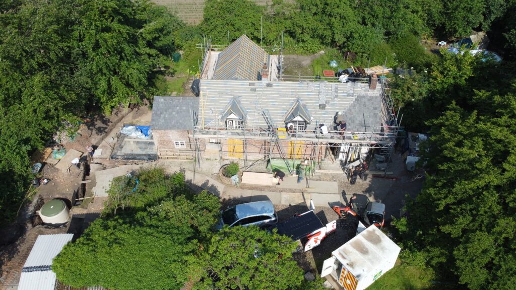 Aerial view of a house under renovation with scaffolding on the roof, construction materials around, workers on site, several vehicles parked nearby, and trees surrounding the area.