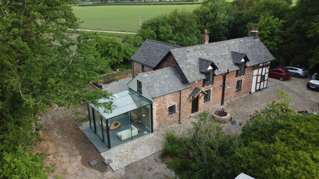 Aerial view of a traditional brick house with a modern glass extension, surrounded by trees and fields. There are cars parked nearby and a stone path leads to the front entrance.