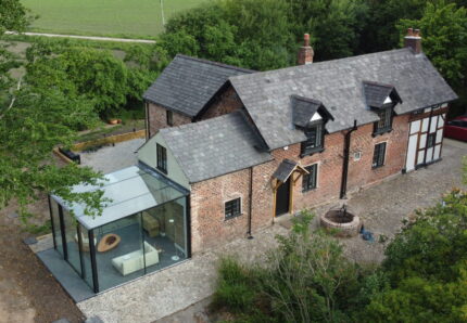 Aerial view of a brick house with a modern glass extension, surrounded by trees and fields, with several parked cars and a gravel driveway.