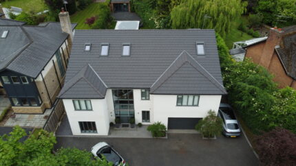 Aerial view of a modern two-story house with a gray tile roof, white walls, large front windows, and a double garage. Two cars are parked in the driveway, and the property is surrounded by greenery and neighboring houses.