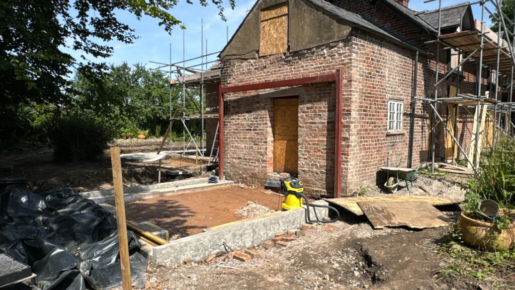 A brick house under construction with scaffolding, boarded windows and doors, and a new concrete foundation in the foreground. Construction materials and tools are scattered around the site.