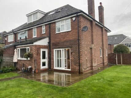 A two-story brick house with several windows, white-framed patio doors, and a small dormer on the top. The backyard features a green lawn, a wooden fence, and a wet stone patio with garden tools and a white umbrella. It's an overcast day.
