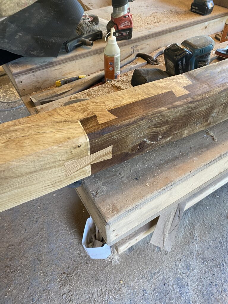 A close-up of a wooden beam with intricate joinery, showing light and dark wood pieces fitted together. Tools, wood glue, and sawdust are visible on a workbench in the background, indicating active woodworking.