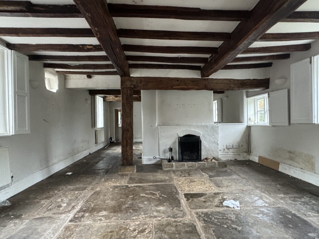 A rustic, empty room with exposed wooden ceiling beams, stone floor, white walls, a central fireplace, and several small windows with open shutters that let in natural light.