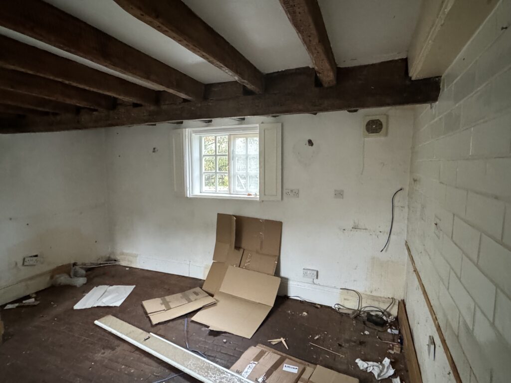 An empty, rundown room with exposed wooden ceiling beams, a small window, dirty white walls, and scattered cardboard and debris on a worn wooden floor.