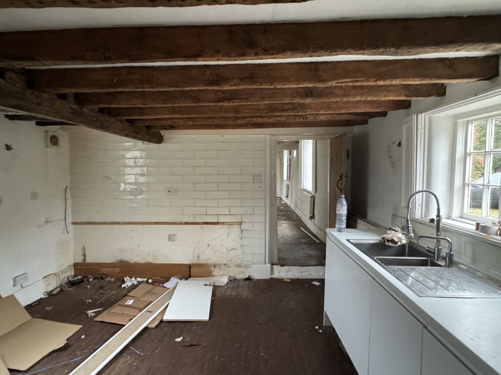 An old, partially renovated kitchen with exposed wooden ceiling beams, white tile walls, a modern sink, scattered debris, and a window bringing light into the worn space. An open doorway leads to another room.