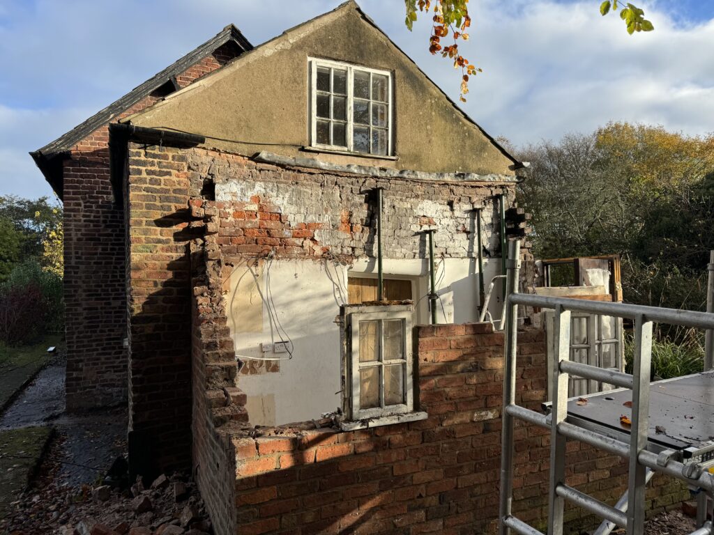 Partially demolished brick house with exposed interior walls and two windows, surrounded by autumn trees and construction scaffolding under a partly cloudy sky.