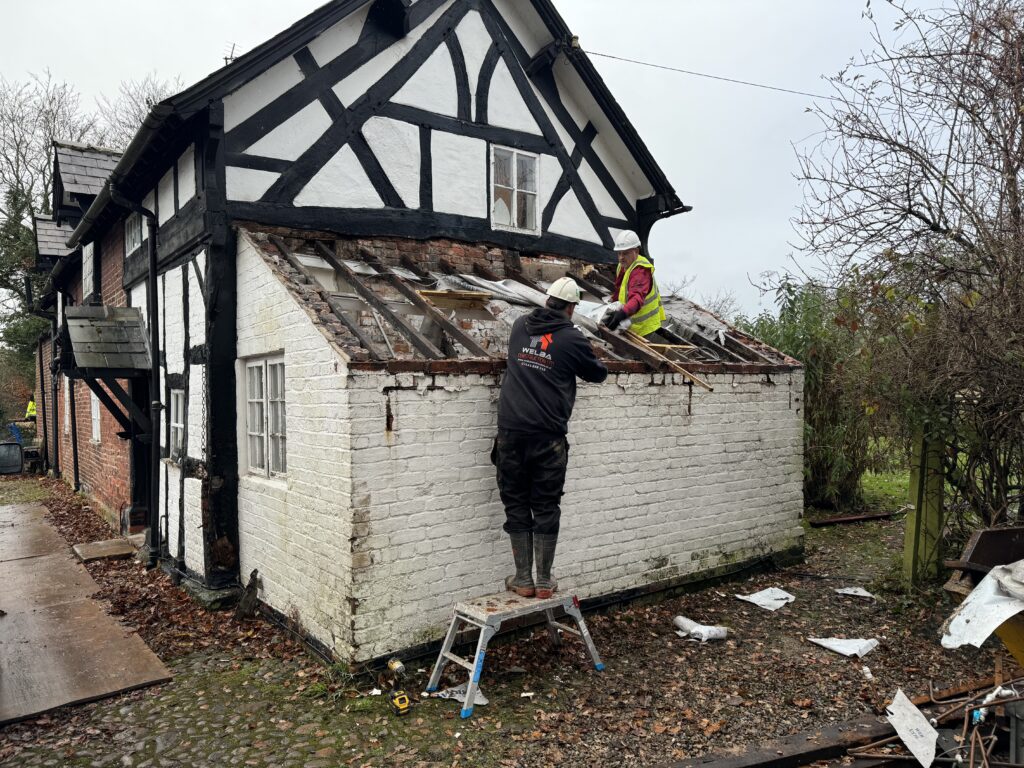 Two people in work gear remove the roof of a white brick building with exposed beams. One stands on a stepladder, the other sits on the roof. Debris and tools are scattered on the ground. Trees and another worker are in the background.