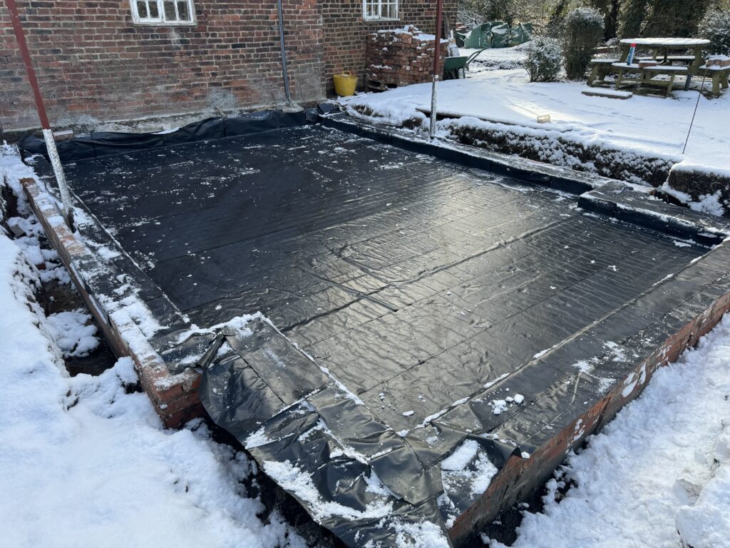 A snow-covered construction site shows a brick foundation with a large black plastic sheet covering the ground. Snow surrounds the area, and a brick building and picnic table are visible in the background.