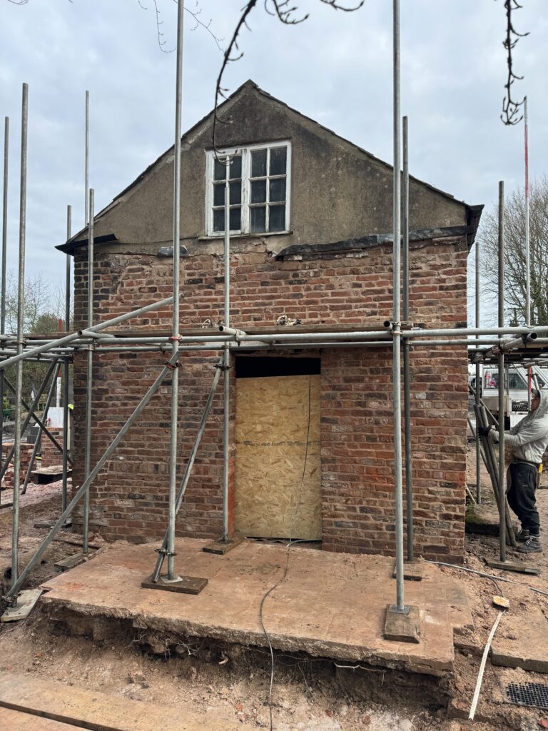 A small brick building under renovation, surrounded by scaffolding. The door is boarded up with plywood, and the front step is wooden. The building has an old window above the door and weathered exterior walls.