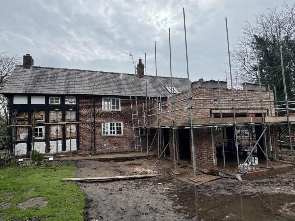 A brick house under renovation with scaffolding and partially exposed timber framing. An extension is being built on the right, with open brickwork and metal poles. The ground is muddy and construction materials are visible.