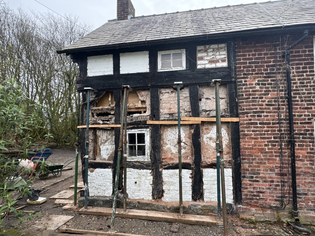 A partially restored brick and timber house with exposed wooden beams, damaged plaster, and support posts. A small white window remains intact amid the repairs. Vegetation and tools are visible nearby.