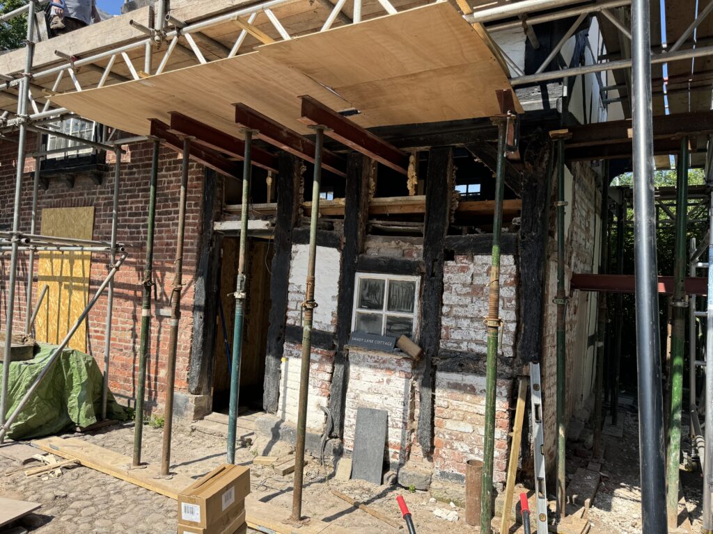 A partially demolished brick and timber building under restoration, supported by metal scaffolding and poles, with wooden boards overhead and construction materials scattered nearby.