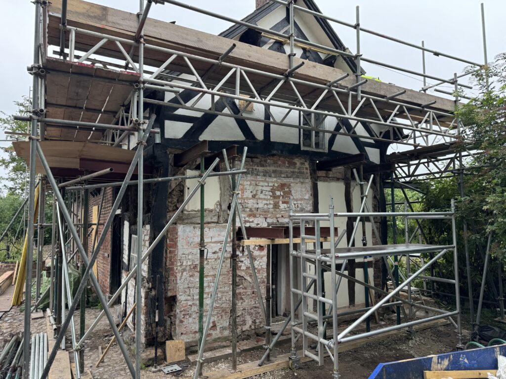 A house under renovation with scaffolding on all sides. The exterior walls are partially stripped, revealing brickwork and timber framing. Construction materials and tools are scattered nearby.