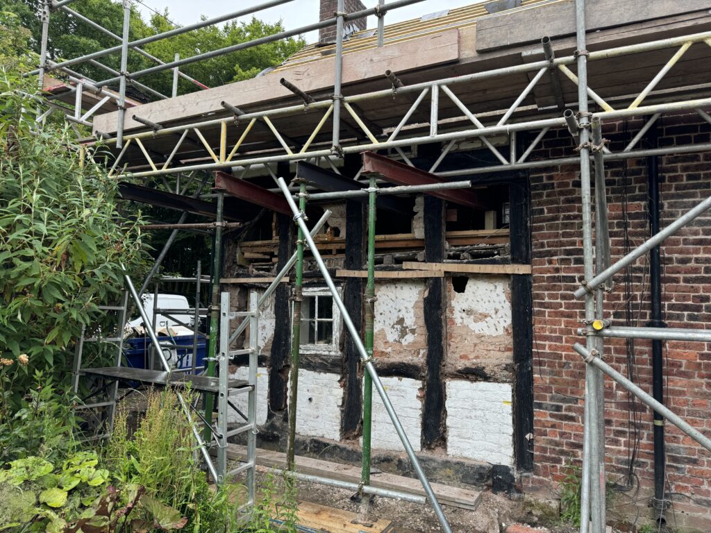 A brick and timber-framed building under renovation, surrounded by scaffolding. The wall is partially exposed, revealing wooden beams and insulation. Vegetation grows in the foreground.