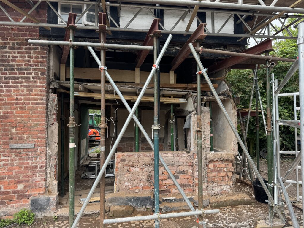 A brick house under renovation with scaffolding and steel supports in place. The front wall is partially demolished, exposing the interior, and construction materials are visible around the site.