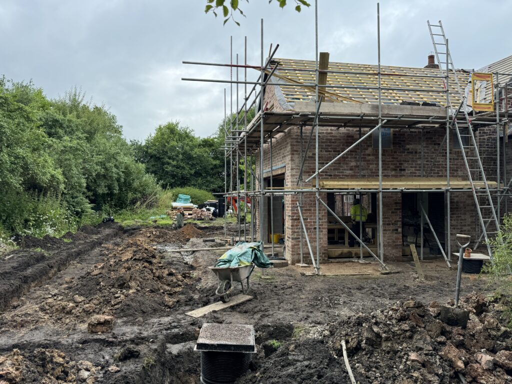 A partially constructed brick house with scaffolding on the roof, surrounded by muddy ground, building materials, and construction equipment, with trees and greenery in the background.