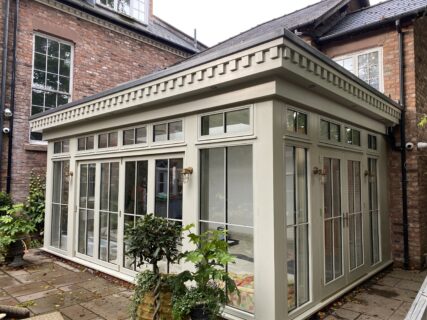 A modern glass conservatory with cream-colored frames is attached to a brick house, surrounded by potted plants on a stone patio. The conservatory has large windows and a patterned trim at the roofline.