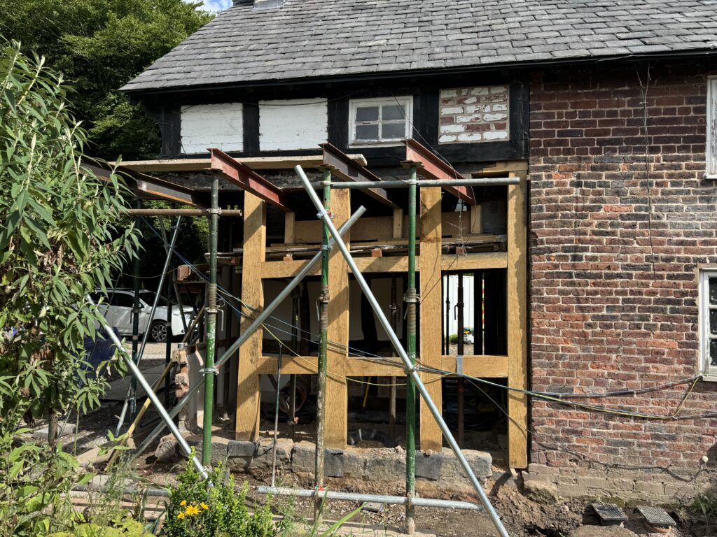 Wooden and metal scaffolding supports a partially renovated brick house with exposed beams and an unfinished wall. Construction materials and a small garden are visible in the foreground.