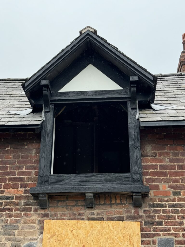 A brick building features a roof dormer with dark wood trim. The dormer window is missing glass, leaving an open rectangular space. The roof is covered with gray shingles, and a plywood board is visible below.
