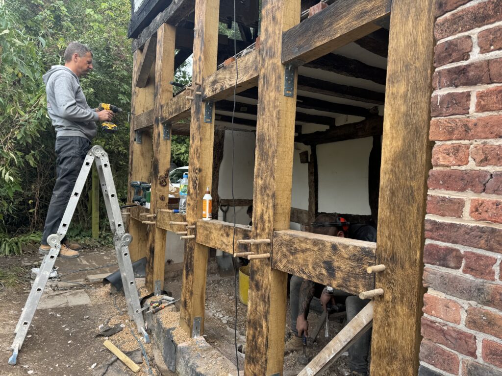 A man stands on a ladder using a tool to work on the timber frame of a partially restored building. Another person works below, surrounded by construction materials, with exposed beams and brick visible.