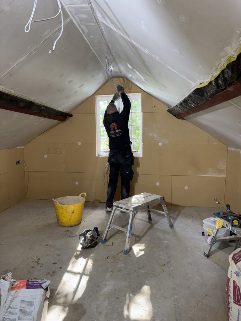 A person stands on a small ladder, working on the ceiling of an unfinished attic room with drywall installed. Construction tools, materials, and a yellow bucket are scattered on the floor. Sunlight shines through a window.