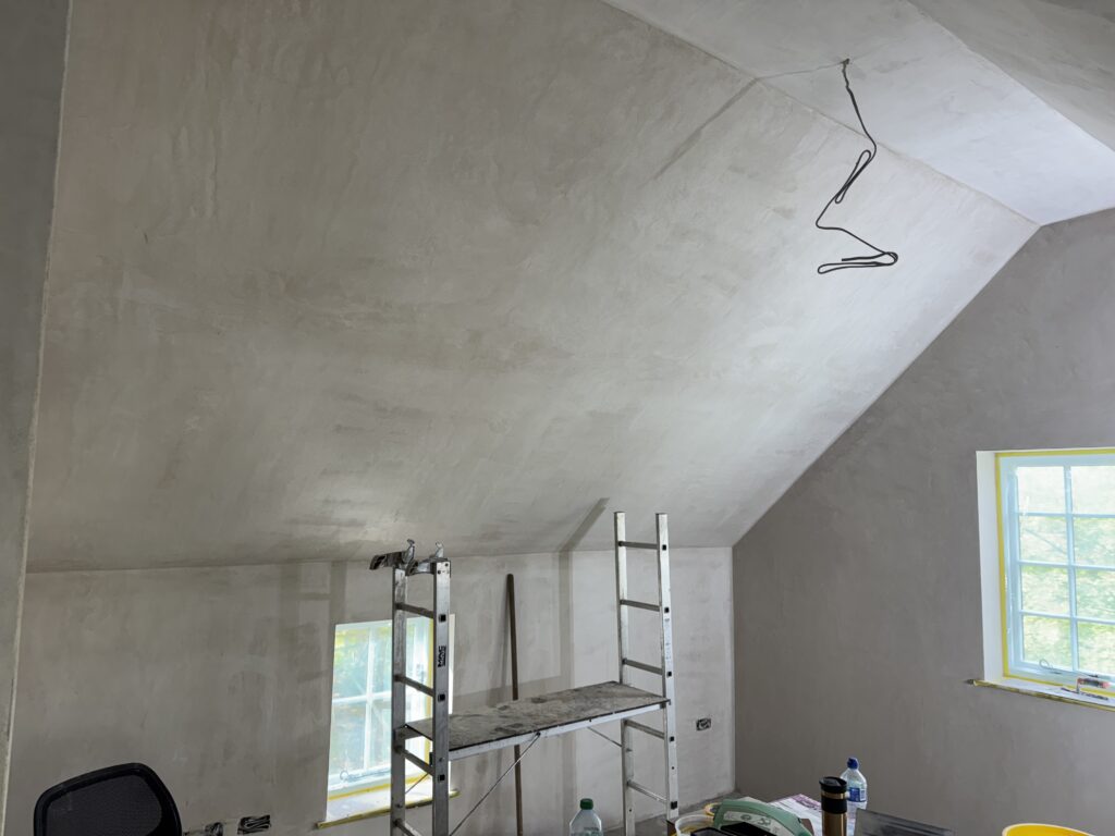 A room with freshly plastered, slanted ceilings and walls. A metal scaffold stands near the wall, and construction materials are scattered below. Sunlight comes through a window with yellow tape on the frame.