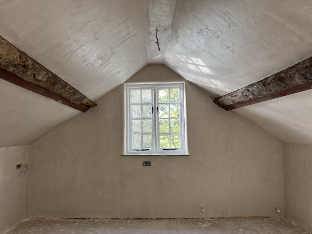 A newly plastered attic room with exposed wooden beams and a central window letting in natural light. The floor is unfinished and the walls are a smooth, light beige color.