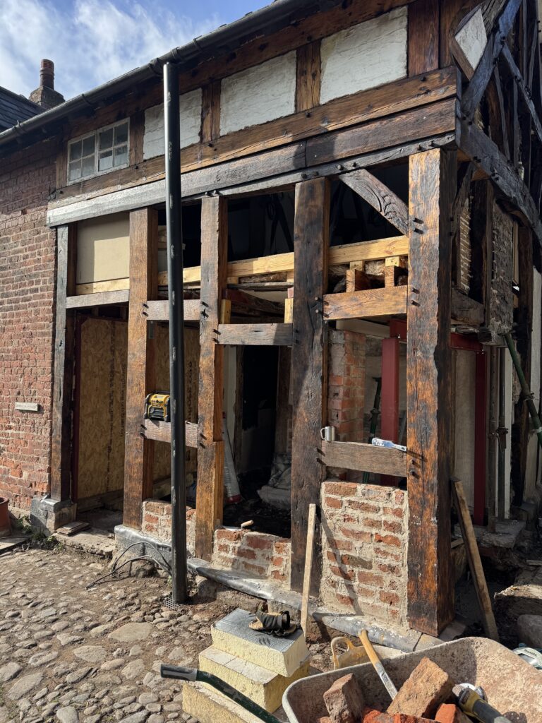 A partially restored timber-framed building with exposed wooden beams and brickwork. Construction tools and materials are scattered outside, and the building’s structure is visible through open areas in the wall.