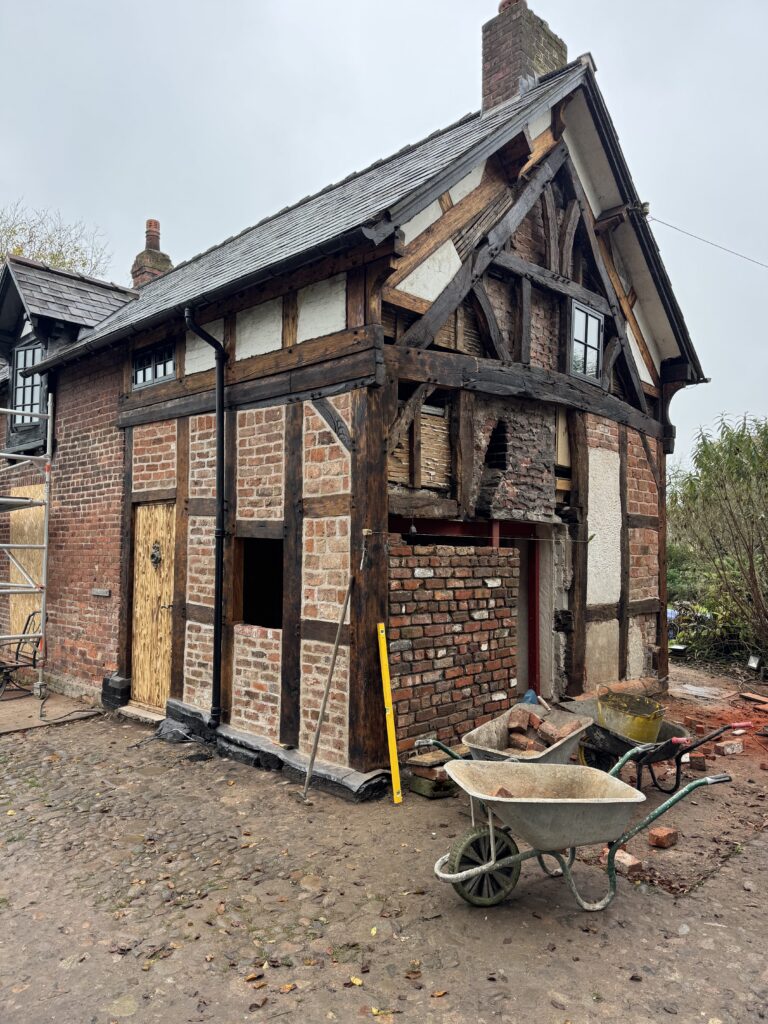 A half-timbered house under renovation with exposed wooden beams, bricks, and a boarded-up door; construction materials and a wheelbarrow are visible in front of the building on a cloudy day.
