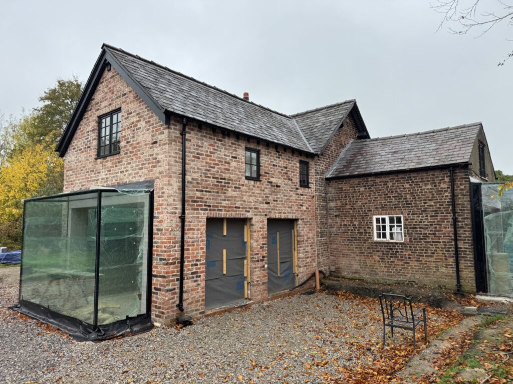 A brick house with black-framed windows and slate roof sits on a gravel area. The house has two sections, one taller than the other, and a glass structure is attached to the left side. Autumn leaves cover the ground.