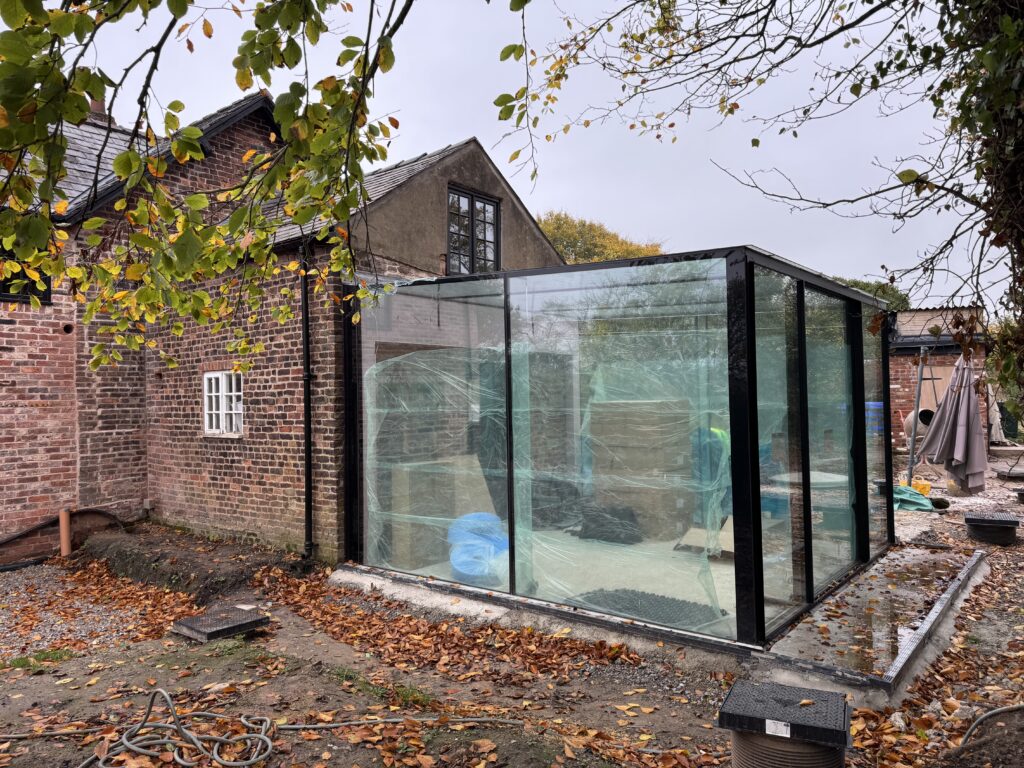 A modern glass room extension is attached to an old brick house surrounded by fallen autumn leaves. Construction materials are visible inside the glass structure, and trees with yellow leaves frame the scene.