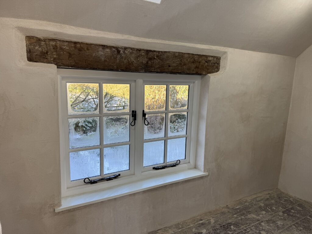 A white-framed window with six panes is set in a plastered wall. An old wooden beam acts as a lintel above the window. The outside view shows frost on the lower window and trees in the background.