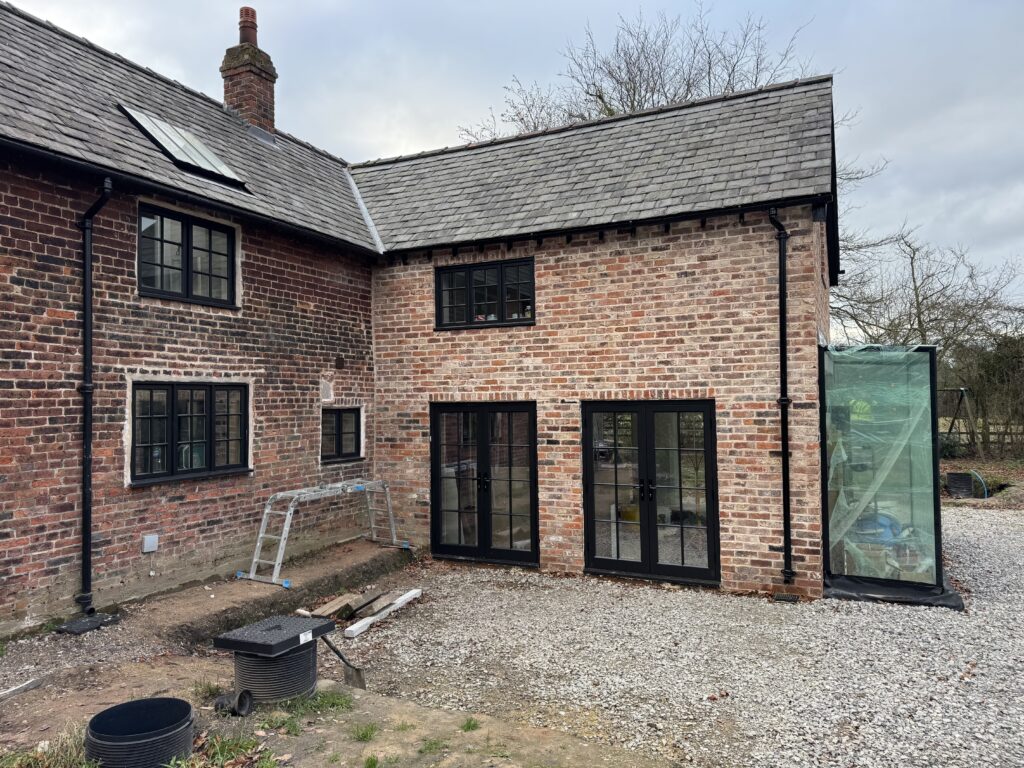 A brick house with black-framed windows and doors, a new extension on the right, construction materials, and a ladder outside. The ground is covered with gravel and dirt, and there are bare trees in the background.