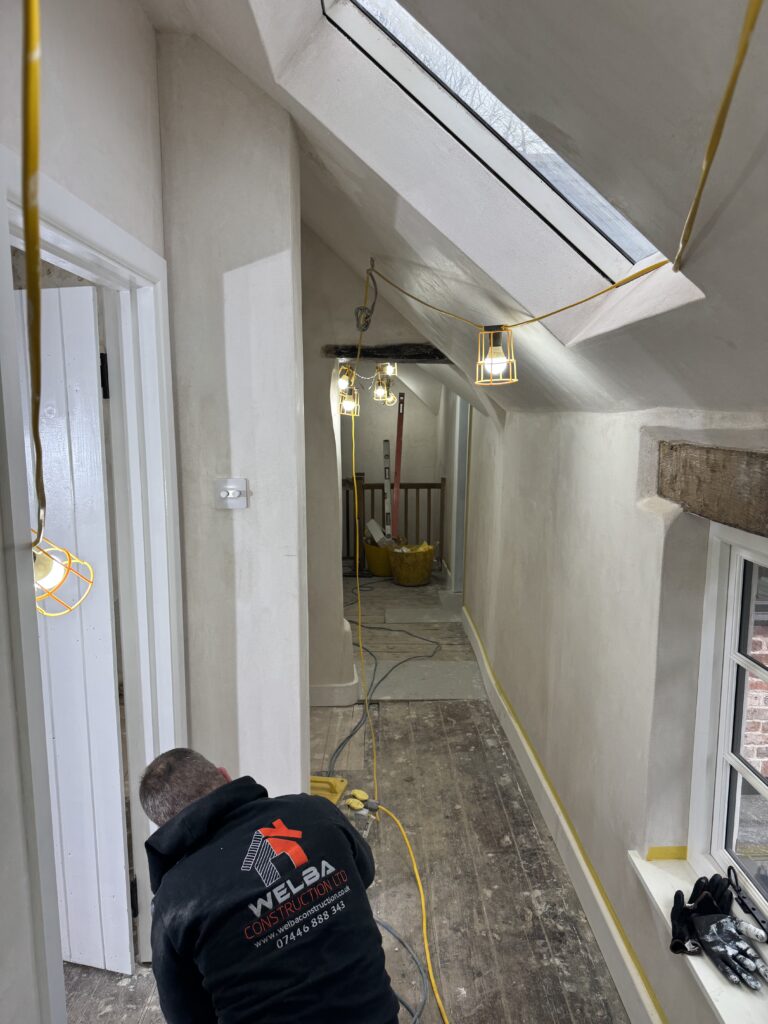 A construction worker kneels in a narrow, freshly plastered hallway with exposed floorboards and temporary work lights hanging from the ceiling. A skylight and windows let in daylight. The worker wears a black hoodie with a company logo.