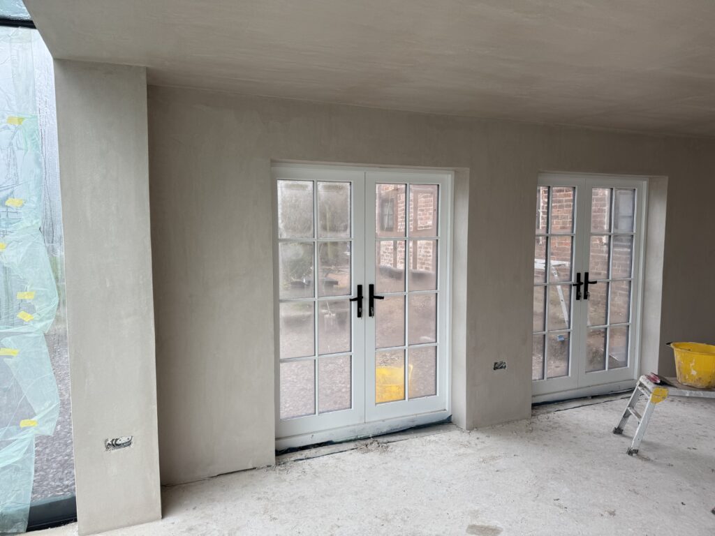 A room under construction with white-framed French doors, unpainted plaster walls, and a concrete floor. A yellow bucket and a step ladder are in the corner. Plastic covers part of a large window on the left.