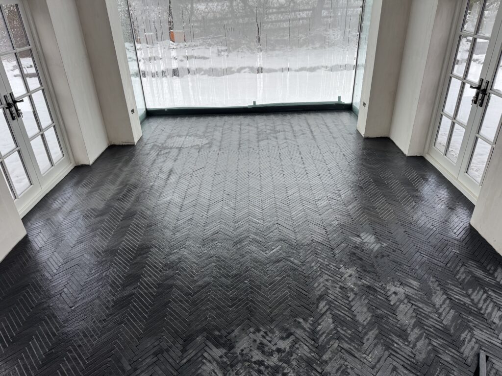 A sunroom with large windows and glass doors on three sides, showing a snowy outdoor scene. The floor features a dark herringbone tile pattern.