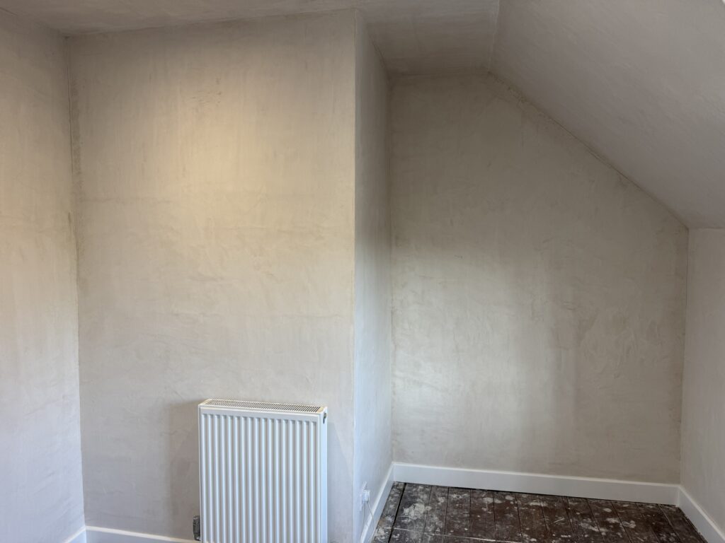 A small, freshly plastered room with cream-colored walls, a white radiator, white skirting boards, and a bare, unfinished floor. The room has a sloped ceiling on the right side.