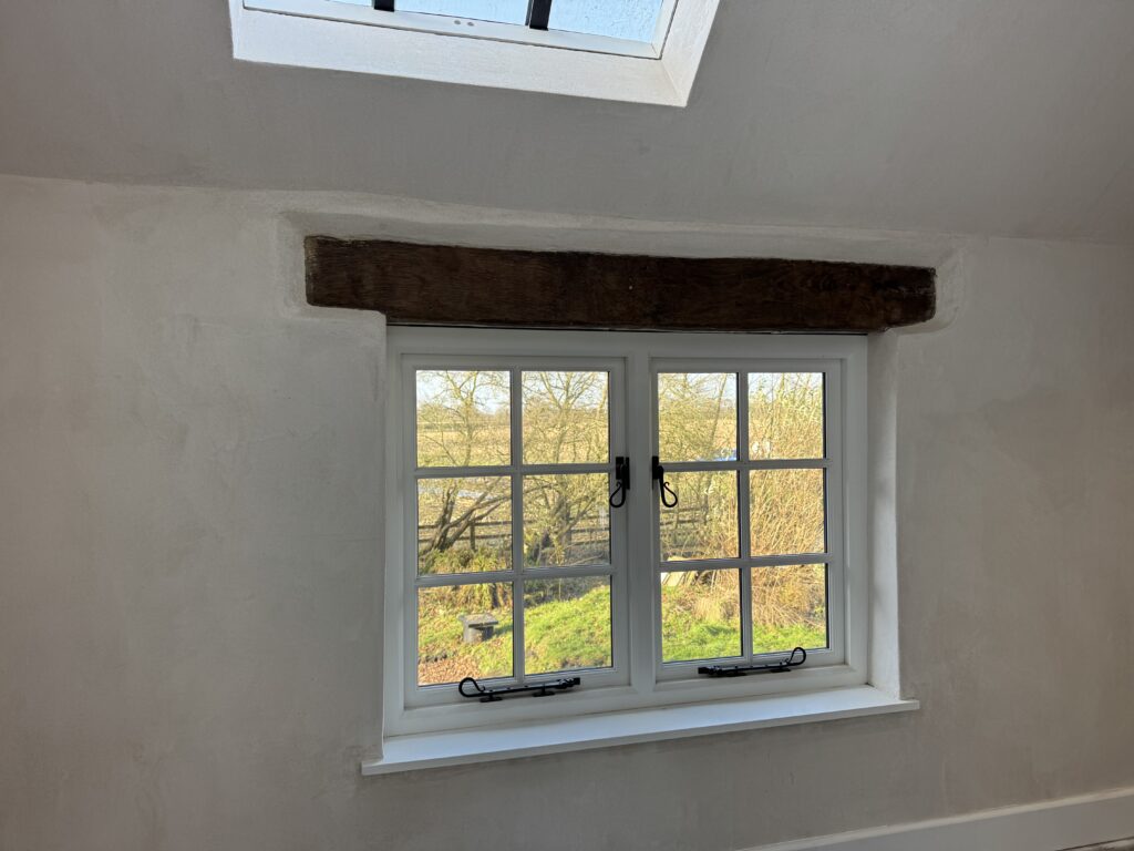 A white-framed window with metal latches set in a plastered wall under a wooden beam, showing a view of bare and leafy trees and green grass outside. A skylight is visible in the ceiling above.