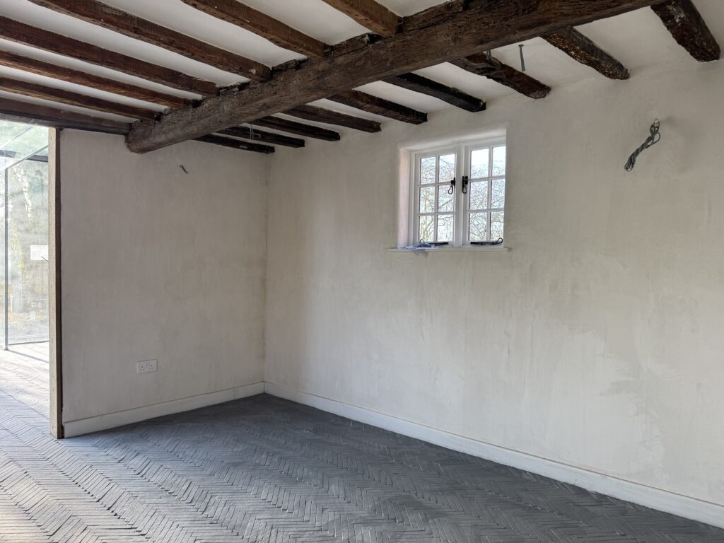 A bright, empty room with a small window, exposed wooden ceiling beams, light-colored plaster walls, and patterned gray tile flooring. Wires stick out from the ceiling and walls, suggesting ongoing renovation.